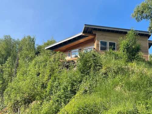 A modern house perched on a hillside, surrounded by lush greenery and under a clear blue sky.