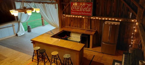 A rustic bar area in a wooden barn, featuring a Coca-Cola sign, stools, and soft lighting.