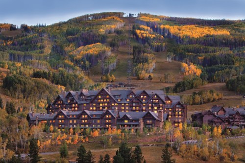 A large lodge nestled in a valley, surrounded by colorful autumn trees and a ski slope in the background.