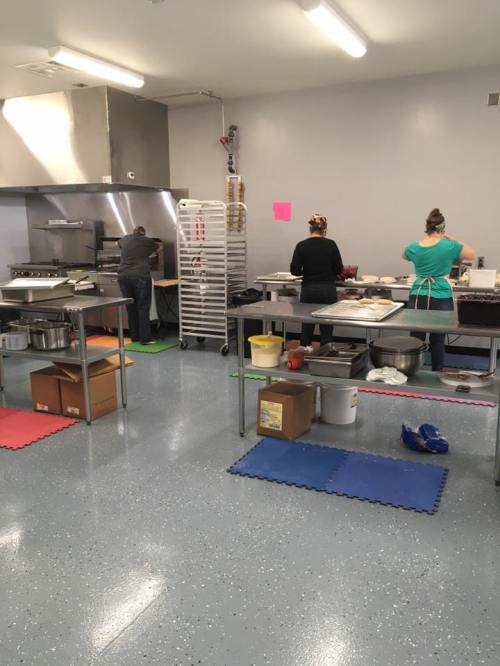 Two people working in a commercial kitchen, preparing food at stainless steel tables with various equipment and supplies.