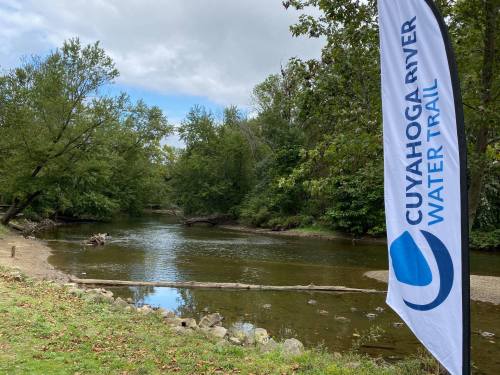 A serene river scene with trees lining the banks and a flag for the Cuyahoga River Water Trail in the foreground.