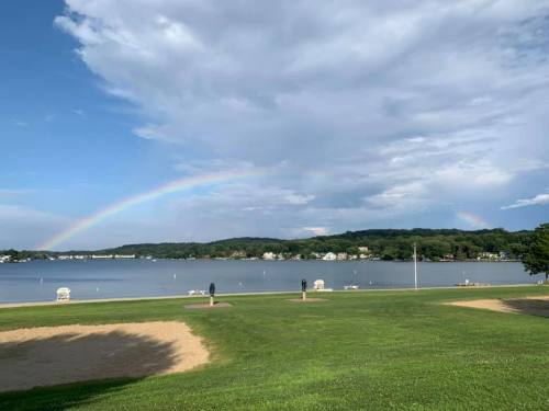 A serene lakeside view with a rainbow arching over the water, surrounded by green grass and distant houses.