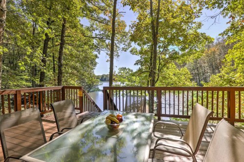 A serene deck overlooking a lake, surrounded by lush trees, with a table set for dining and a bowl of fruit.
