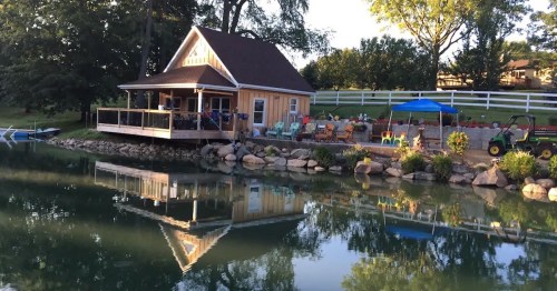 A cozy lakeside cabin with a deck, surrounded by greenery and reflecting in the calm water. Colorful chairs are visible.