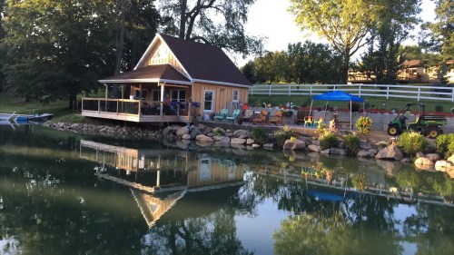 A serene lakeside scene featuring a wooden house, patio furniture, and reflections in the calm water.