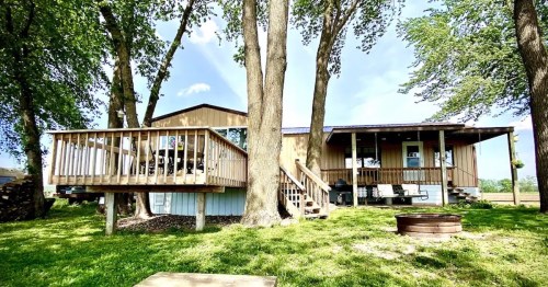 A wooden house with a deck surrounded by trees, featuring a fire pit in the yard and a clear blue sky above.