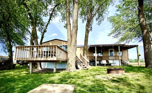 A cozy house surrounded by trees, featuring a deck and outdoor seating area, set in a green landscape.