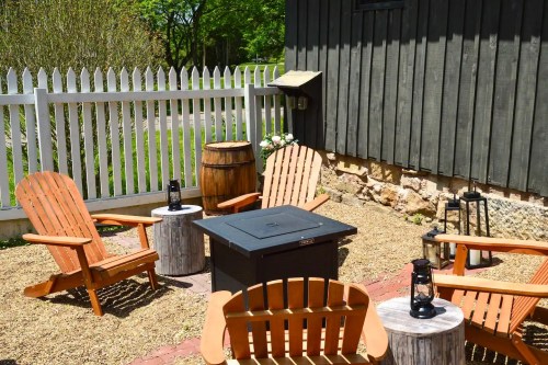 A cozy outdoor seating area with wooden chairs, a fire pit, and lanterns, surrounded by a white picket fence.