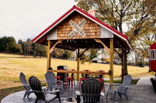 A cozy outdoor gazebo with seating, surrounded by trees and a grassy field, featuring a railroad crossing sign.