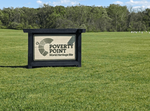 Sign for Poverty Point World Heritage Site in a grassy field, surrounded by trees under a clear blue sky.