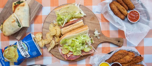 A wooden platter with sandwiches, chips, and fried items, alongside a can of chips on a checkered tablecloth.