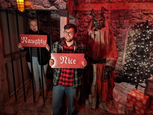 A man holds a "Nice" sign while a woman in a cell holds a "Naughty" sign, with a festive creature nearby.