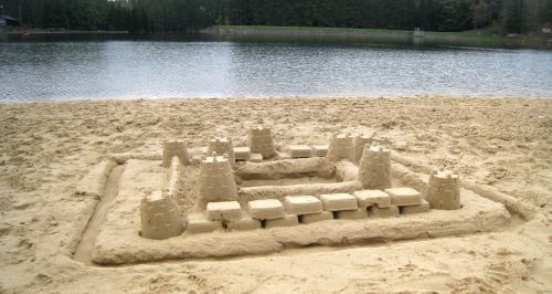 A sandcastle with towers and a wall, built on a sandy beach near a calm lake surrounded by trees.