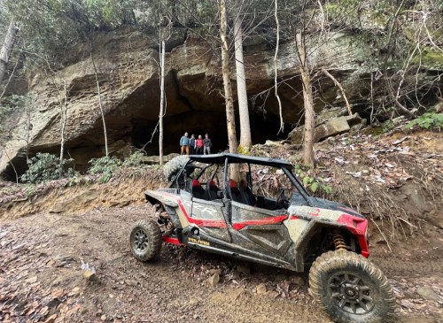 An off-road vehicle parked near a cave entrance, with three people standing in the background among trees and rocky terrain.