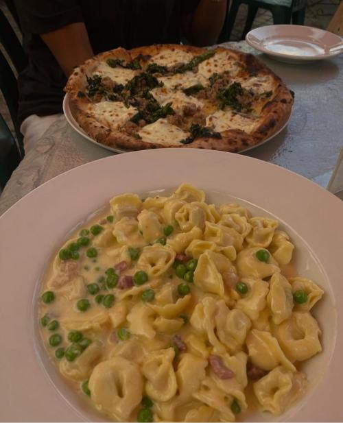 A plate of creamy tortellini with peas in the foreground and a pizza with cheese and greens in the background.