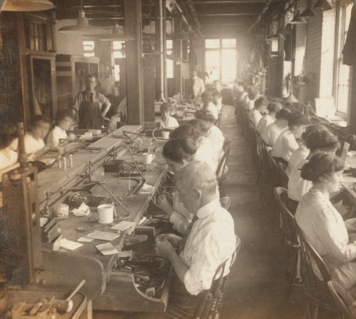 A historic photo of a factory interior with workers at benches, focused on tasks in a well-lit space.