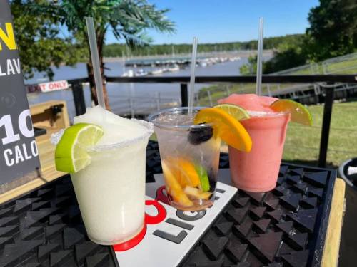 Three colorful cocktails sit on a table, with a scenic view of a marina and trees in the background.