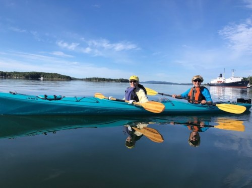 Two people in kayaks on calm water, surrounded by trees and a clear blue sky, with reflections visible in the water.