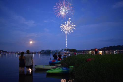 People watch fireworks over a lake while standing near kayaks at dusk, with a full moon in the background.