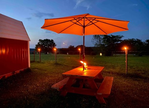 A red picnic table with an umbrella, surrounded by torches, set in a grassy field at dusk.