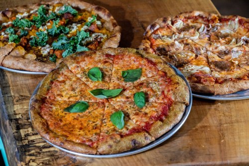 Three pizzas on wooden table: one with greens, one with meat and cheese, and a margherita topped with basil leaves.