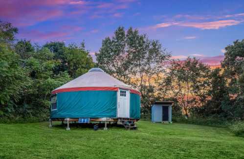 A colorful yurt sits on a grassy area surrounded by trees, with a vibrant sunset sky in the background.