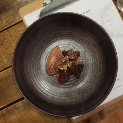 A dark bowl containing a small serving of chocolate dessert topped with crunchy bits, placed on a wooden table.