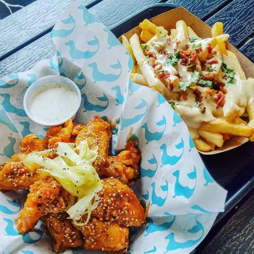 A tray with crispy chicken wings topped with sesame seeds and pickled onions, alongside loaded fries with cheese and bacon.