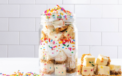 A jar filled with layered dessert squares, whipped cream, and colorful sprinkles, with some squares scattered nearby.