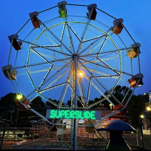 A brightly lit Ferris wheel at dusk, with a sign reading "SUPERSLIDE" in front, surrounded by a festive atmosphere.