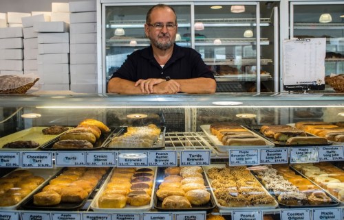 A man stands behind a display case filled with various donuts in a bakery.