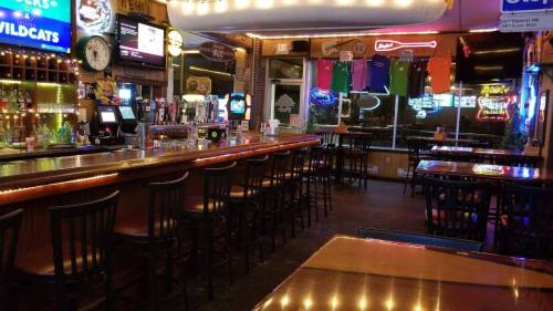 A dimly lit bar with empty stools, neon signs, and a view of the street through large windows.