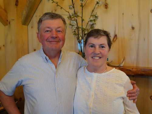 A smiling man and woman stand together in a cozy wooden setting, with flowers in the background.