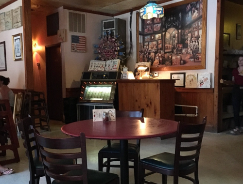 Cozy restaurant interior with a red table, wooden chairs, a jukebox, and a mural on the wall.