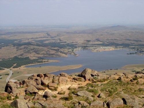 A panoramic view of rolling hills and a lake, with rocky terrain in the foreground and a clear sky above.