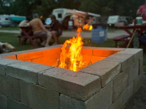 A cozy campfire burns in a stone fire pit, with people gathered around in a campsite setting.