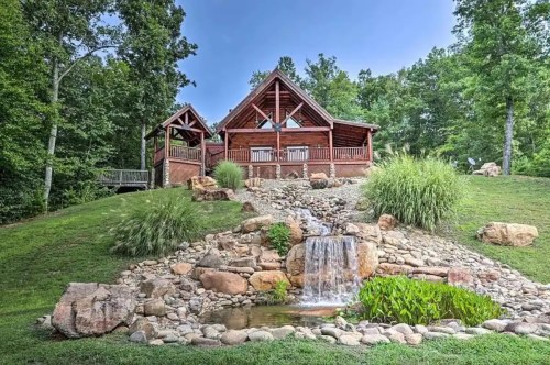 A rustic cabin surrounded by greenery, featuring a small waterfall and landscaped rocks in the foreground.