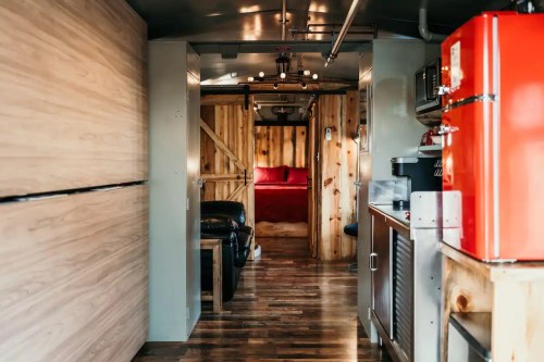 Cozy interior of a tiny home, featuring wooden walls, a red fridge, and a view of a bedroom with a red bedspread.