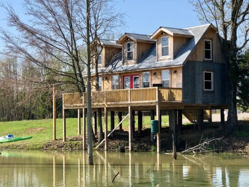 A wooden house on stilts by a calm lake, surrounded by trees and greenery, with a kayak visible nearby.