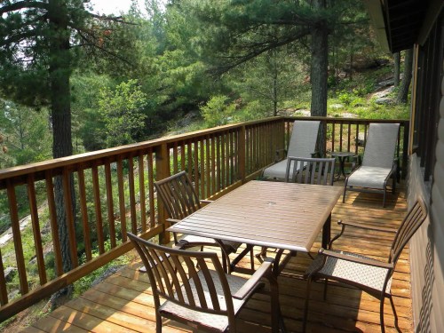 A wooden deck with a table and chairs, surrounded by trees and greenery. Two lounge chairs are also visible.
