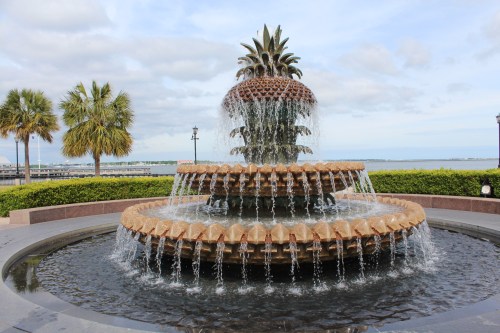 A decorative pineapple fountain with cascading water, surrounded by greenery and a view of the water in the background.