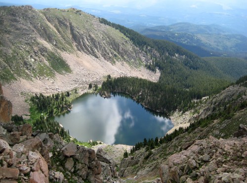 A serene mountain lake surrounded by lush greenery and rocky terrain, reflecting clouds in its calm waters.