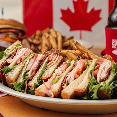 A platter of stacked sandwiches with lettuce, tomato, and turkey, served with crispy fries and a Canadian flag in the background.