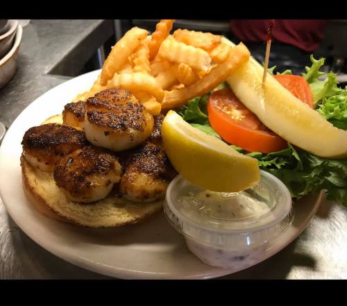 A plate featuring grilled scallops on a bun, with fries, lettuce, tomato, and a lemon wedge, plus a side of dipping sauce.