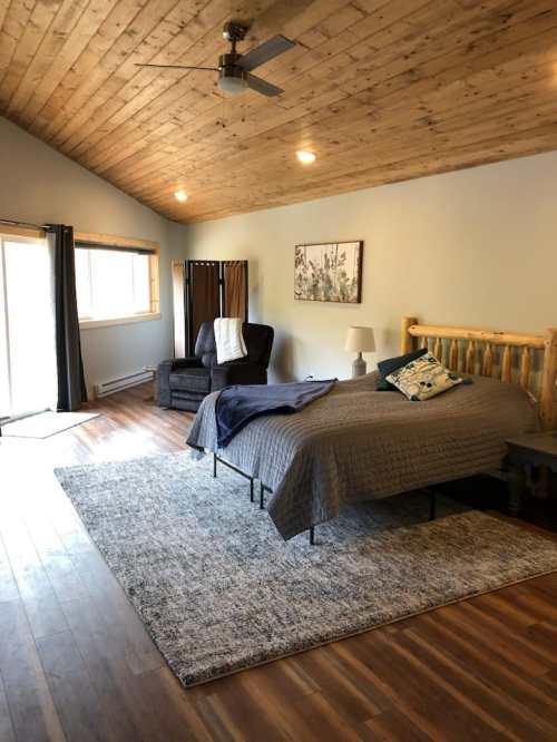 Cozy bedroom with wooden ceiling, a bed, armchair, and large window, featuring a rug and soft lighting.