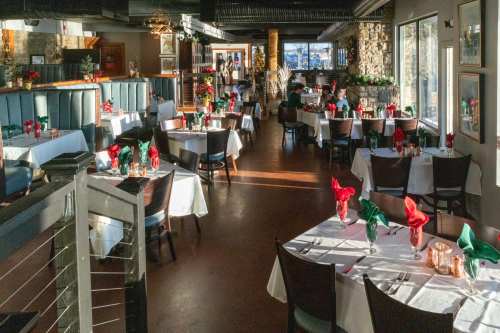 A spacious restaurant with tables set for dining, featuring red and green napkins, and warm sunlight streaming in.
