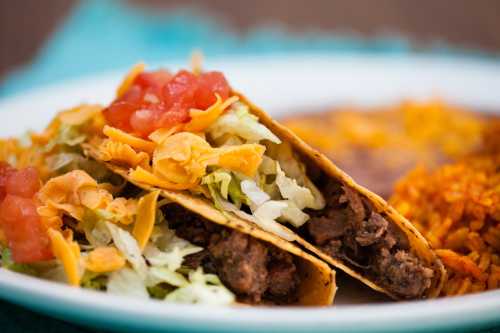 Two tacos filled with seasoned beef, lettuce, cheese, and diced tomatoes, served with rice and beans on the side.