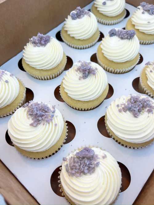 A box of vanilla cupcakes with swirled white frosting and purple crystal sprinkles on top.