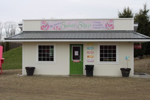 A small, white sweet shop with a green door, featuring signs for fudge, candies, and sweet treats.