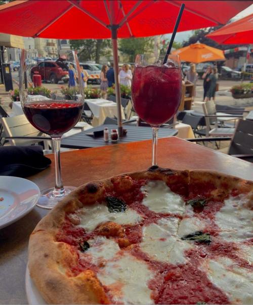 A close-up of a margherita pizza with two drinks on a table, outdoor dining scene with umbrellas in the background.
